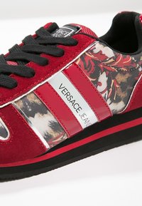 Baskets rouges et noires avec motif floral, lacets noirs et panneau blanc avec le texte « Versace Jeans » sur le côté.