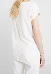 Camiseta blanca de manga corta hecha de un tejido texturizado. Presenta puños enrollados y un dobladillo posterior ligeramente más largo.