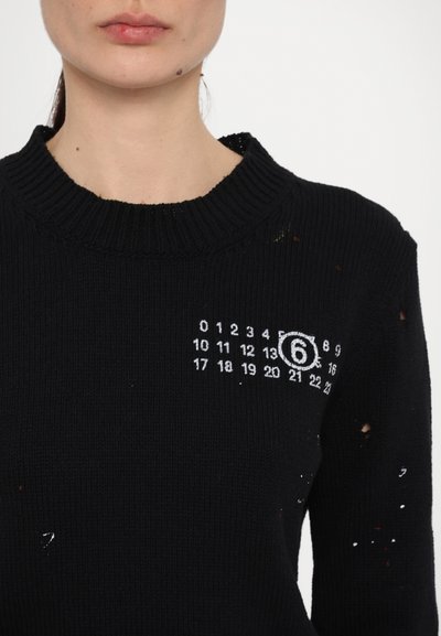 Svart stickad tröja med en bred ribbad krage, med hål och ett distinkt vitt nummertryckdesign på framsidan.