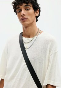 White knit shirt with short sleeves and button detail, paired with layered necklaces and a black crossbody strap. Neutral background.