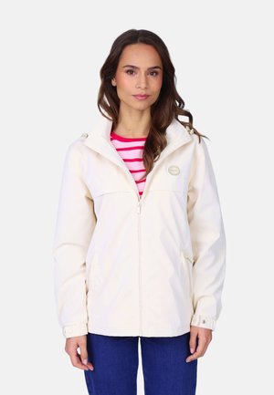 Jeune femme portant une veste zippée écrue sur un haut rayé rose et blanc, associée à un jean bleu, debout devant un fond uni.