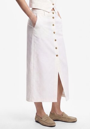 Donna che indossa una gonna midi bianca con bottoni frontali, tasche e mocassini beige, in piedi davanti a uno sfondo semplice.