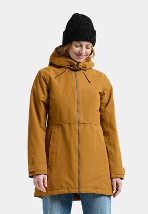 Kvinna som bär en senapsgul huva med dragkedja framtill och en svart stickad mössa, ler och tittar lite nedåt.