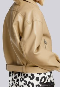 Giacca in pelle marrone con un design strutturato, maniche oversized e una tasca con cerniera, abbinata a una camicia bianca e nera con stampa leopardata sotto.
