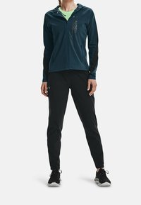 Sudadera con cremallera en azul verdoso con mangas negras, que presenta un logo. Combinada con pantalones negros ajustados y zapatillas deportivas negras. Textura suave, corte atlético.