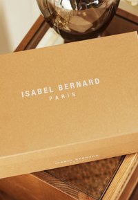 Kartonnetje met een gladde textuur, lichtbruine kleur en witte tekst met de opdruk "ISABEL BERNARD PARIS," geplaatst op een houten oppervlak.