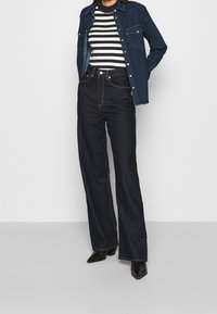 Jean évasé en denim foncé associé à un haut côtelé à rayures noires et blanches sous une veste en denim bleu. Des bottines noires à bout pointu complètent le look.