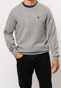 Grå stickad tröja med rund hals och ribbade manschetter, med en liten marinblå logotyp på bröstet. Matchad med svarta byxor.