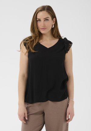 Woman with long brown hair wearing a black sleeveless top with lace detail on one shoulder and taupe pants standing against white background.