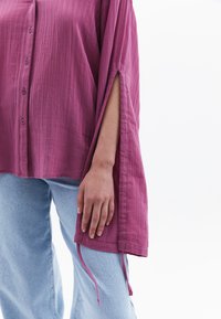 Camicia button-up in un tessuto rosa profondo, con maniche lunghe e spacchi laterali, dotata di lacci all'orlo. Indossata con jeans azzurri chiari.