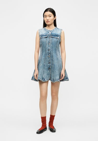 Γυναίκα που φοράει αμάνικο ανοιχτό μπλε τζιν φόρεμα με κουμπιά στο μπροστινό μέρος, κόκκινες κάλτσες και μαύρα πλατφόρμες, στέκεται μπροστά σε λευκό φόντο.