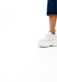 Kāju valkājot īsās zilās džinsa bikses un baltu masīvu platformas sporta apavu uz baltas grīdas ar vienkāršu baltu fonu.