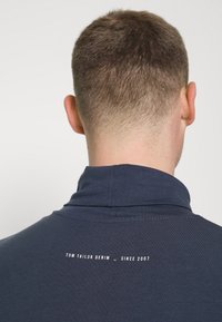 Tummansininen t-paita, jossa on korkea kaulus ja valkoista tekstiä selässä, joka kuuluu "TOM TAILOR DENIM - SINCE 2007." Sujuva pinta.