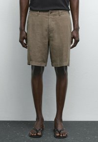 Linnebyxor i en dämpad brun färg, med rak passform, framknappning och sidofickor. Bärs med svarta flip-flops.