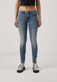 Ljusblå skinny jeans med blekningar och whiskeringdetaljer, femficksdesign och knäppning med knapp, tillsammans med glänsande silverfärgade skor.