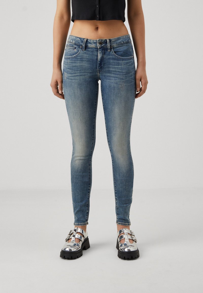 Ljusblå skinny jeans med blekningar och whiskeringdetaljer, femficksdesign och knäppning med knapp, tillsammans med glänsande silverfärgade skor.