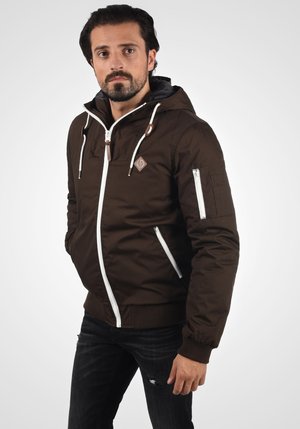 Bruine gewatteerde jas met een capuchon, witte rits en trekkoorden, twee ritszakken aan de voorkant en een patch op de borst; gecombineerd met donkere jeans.