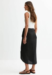 Jupe midi noire avec une finition texturée, associée à un débardeur blanc à côtes et des sandales marron avec des accents à boucles.