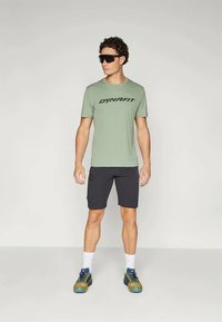 Men's lightweight green DYNAFIT t-shirt with black logo, paired with black shorts, white socks, and multi-colored athletic shoes.