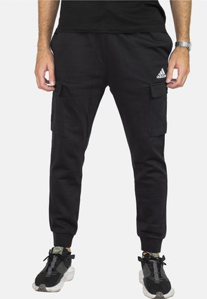 Mann trägt schwarze Adidas Cargo-Jogginghosen und schwarze Turnschuhe mit weißen Sohlen, steht vor weißem Hintergrund.