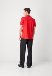Camisa polo roja con cuello, mangas cortas y dobladillo recto, combinada con pantalones negros y zapatos negros, vista desde la parte trasera.
