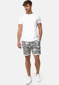 Camiseta blanca de algodón para hombre combinada con pantalones cortos grises con patrones de hojas tropicales blancas y puños enrollados. Zapatillas deportivas blancas completan el look.