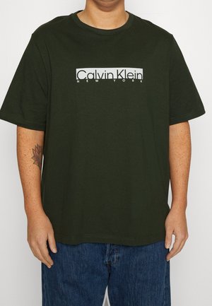 Osoba ubrana w ciemnozieloną koszulkę Calvin Klein z białym logo i niebieskie dżinsy, z widocznym tatuażem na lewym przedramieniu.