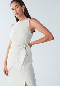 Lichtbeige linnenjurk zonder mouwen, met een knoopdetail in de taille en een split aan de zijkant voor extra beweging en stijl.