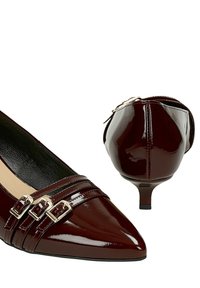 Donkerbruine lakleren damesschoenen met kittenhakken, puntige neuzen en drie kleine zilveren gespsluitingen aan de voorkant.