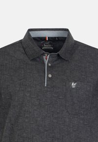 Polo gris foncé en tissu texturé, avec un col, trois boutons, une poche poitrine et un accent logo. Présente des coutures contrastantes.