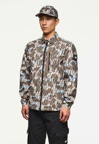 Uomo vestito con giacca a fantasia mimetica e berretto coordinato, con pantaloni cargo neri, in piedi davanti a uno sfondo bianco semplice.