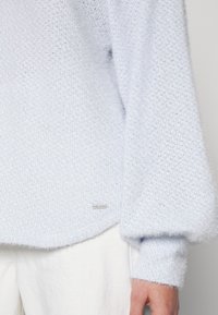 Ljusblå, texturerad stickad tröja med lös passform; har en etikett på fållen och ribbade ärmslut. Mjuk, fluffig look.