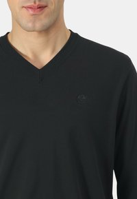 Maglione nero a scollo a V realizzato in tessuto a coste, con un piccolo logo sul petto. La texture appare liscia e leggermente elastica.