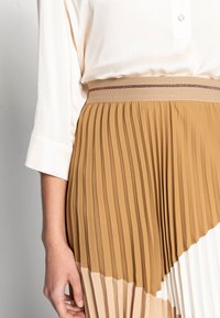 Skládaná sukně v odstínech tan, krémové a bílé. Má texturovaný design s širokým elastickým pasem a subtilním zlatým akcentem.
