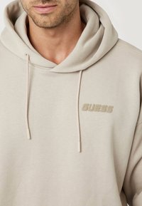 Šviesiai smėlinės spalvos džemperis su reguliuojama raišteliu ir minkšta tekstūra. Krūtinėje turi iškilų "GUESS" logotipą.