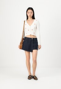 Cardigan bianco a coste con accenti a forma di cuore rossi, abbinato a una gonna avvolgente in denim blu scuro e sandali neri con cinturini. Borsa a spalla marrone inclusa.
