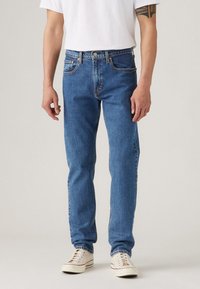 Homme portant un jean bleu droit, des baskets blanches et un t-shirt blanc, debout devant un fond uni.
