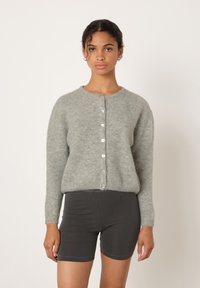 Žena s kudrnatými vlasy, která nosí světle šedý kardigan na knoflíky a tmavě šedé přiléhavé šortky, na jednoduchém světle pozadí.