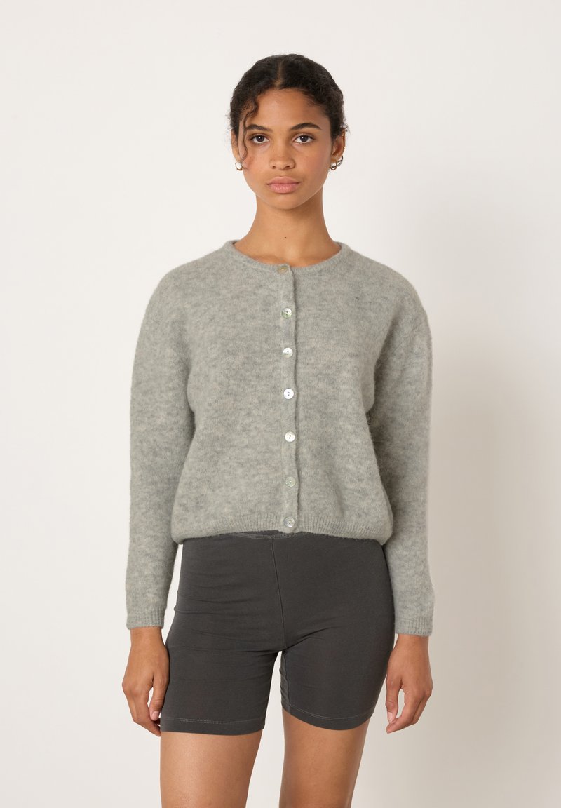 Žena s kudrnatými vlasy, která nosí světle šedý kardigan na knoflíky a tmavě šedé přiléhavé šortky, na jednoduchém světle pozadí.