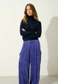 Maglione a collo alto blu navy abbinato a pantaloni in corduroy viola a gamba larga con tasche laterali e texture a righe verticali.