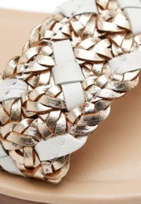 Tira de sandalia de cuero blanco con intrincado trenzado metálico dorado. Presenta una plantilla suave en color beige y un diseño minimalista.