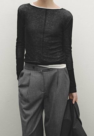 Personne portant un haut noir transparent à manches longues sur une chemise blanche, un pantalon plissé gris, tenant une veste sombre à fines rayures dans une main.