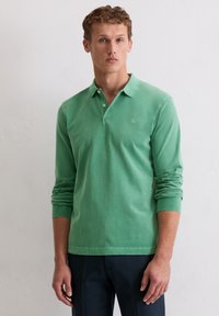 Ein Mann trägt ein langärmliges grünes Poloshirt und dunkle Hosen, steht vor einer weißen Wand, schaut nach vorne mit einem neutralen Ausdruck.