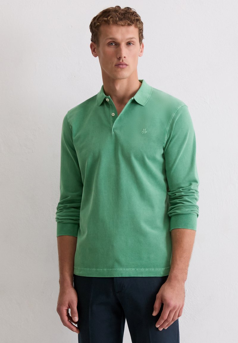 Ein Mann trägt ein langärmliges grünes Poloshirt und dunkle Hosen, steht vor einer weißen Wand, schaut nach vorne mit einem neutralen Ausdruck.