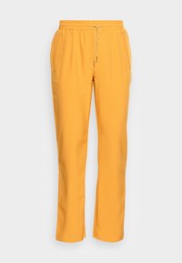 Żółte spodnie o prostych nogawkach z elastycznym pasem i sznurkiem, bocznymi kieszeniami oraz subtelnymi plisami z przodu na jasnoszarym tle.