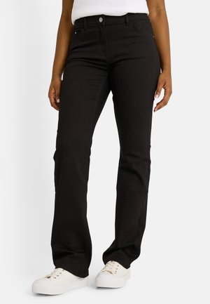 Femme portant un jean bootcut noir et des baskets blanches, se tenant avec les bras détendus contre un fond uni.
