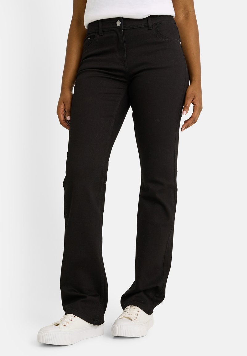 Mujer vestida con jeans bootcut negros y zapatillas blancas, de pie con los brazos relajados contra un fondo sencillo.