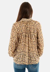 Blusa leggera con motivo floreale in beige, con accenti viola e verdi. Presenta una schiena plissettata e un collo arricciato. Maniche lunghe e a sbuffo.