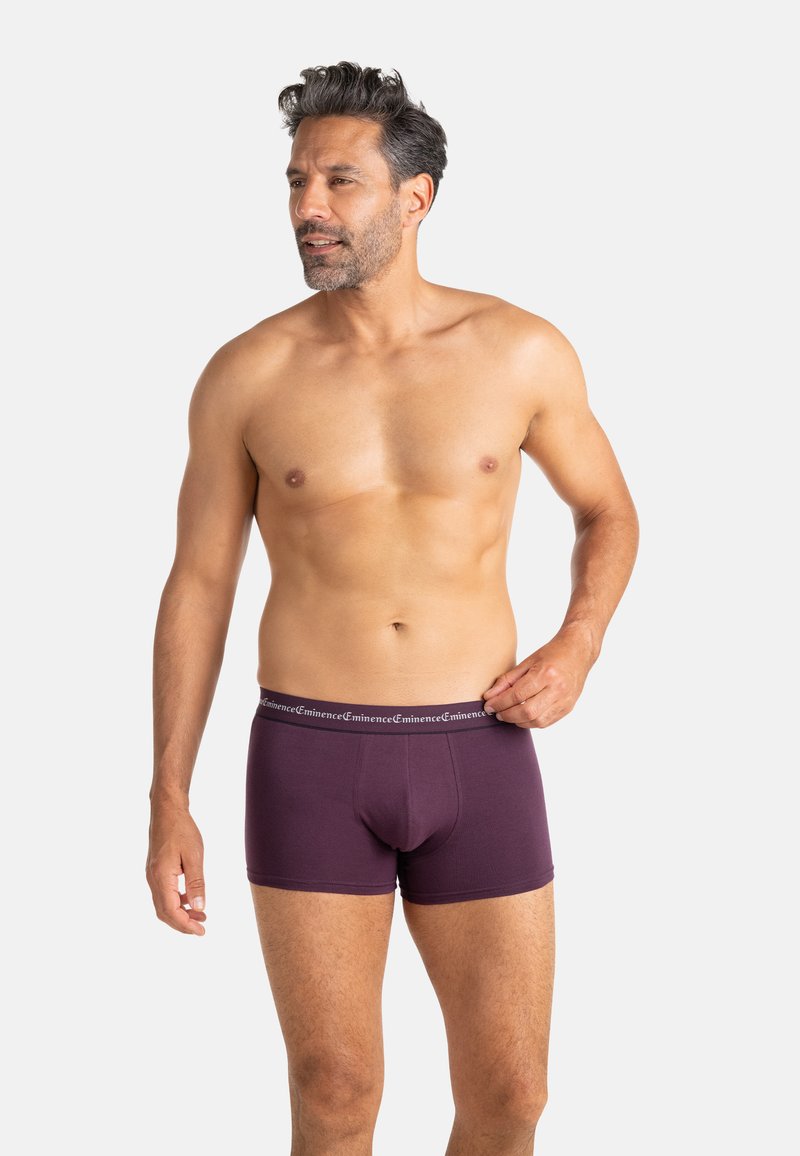 Homme adulte aux cheveux courts et foncés et à la barbe, portant un boxer violet, tenant la ceinture, regardant sur le côté devant un fond uni.