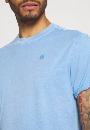 Mann trägt hellblaues T-Shirt mit kleinem Logo auf der Brust und sichtbarem buntem Tattoo am Oberarm vor weißem Hintergrund.